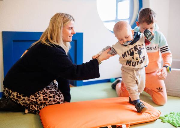 Eine Erzieherin hält einen kleinen Jungen am Arm fest, der auf einem Kissen läuft und ein Handy in einer Hand hält.